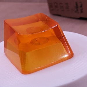 May include: Orange-tinted, square-shaped keycaps, possibly for a keyboard, are displayed on a white cylindrical stand. Three additional keycaps are scattered nearby. A small cardboard box is in the background.