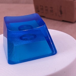 Könnte beinhalten: Vier blaue, transparente Keycaps mit abgerundeter Oberseite und quadratischer Basis. Die Keycaps bestehen aus einem klaren Material, durch das man hindurchsehen kann. Die Keycaps liegen auf einer weißen Oberfläche.