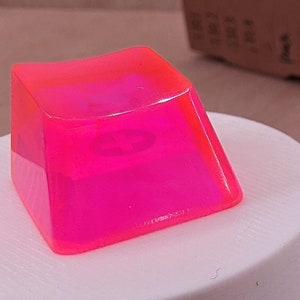 Puede incluir: Cuatro teclas translúcidas de color rosa brillante para un teclado mecánico. Las teclas están hechas de plástico transparente con un tinte rosa. Están sobre una superficie blanca.
