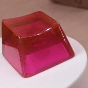 May include: Four pink translucent keycaps for a mechanical keyboard. The keycaps are square shaped and have a slightly curved top. One keycap is sitting on a white surface.