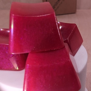 May include: Close-up of four stacked, glossy, red-pink resin keycaps. The keycaps have a slightly curved top and a square base. The resin has a glittery, iridescent effect. The keycaps are on a white surface.