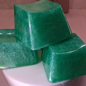May include: Three green resin keycaps with a glittery finish. The keycaps are square shaped and have rounded edges.
