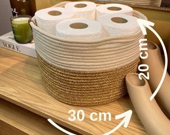Puede incluir: Una cesta de almacenamiento tejida beige y marrón con cuatro rollos de papel higiénico blanco en su interior. La cesta tiene un diámetro de 30 cm y una altura de 20 cm.