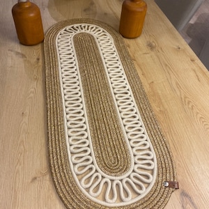 May include: A woven oval table runner with a brown and white braided design. The runner has a brown leather tag on the side.