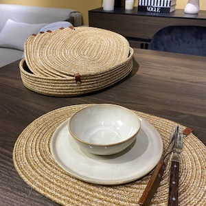May include: A set of oval woven placemats with a white ceramic bowl and plate on top. The placemats are made of natural fibers and have a light brown color. A wooden fork and knife are placed on the placemat.