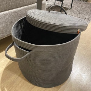 Puede incluir: Una cesta de almacenamiento de cuerda gris grande con tapa. La cesta tiene dos asas en el lateral y una tapa tejida.