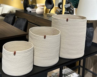 May include: Three white rope baskets of varying sizes, with brown leather handles, are displayed on a black metal shelf. The baskets are arranged in a descending size order, with the largest basket at the top and the smallest basket at the bottom. The shelf is made of black metal and has a glass top.