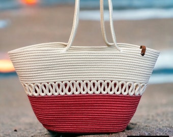 Pode incluir: Uma bolsa de mão de corda branca e rosa com uma alça de corda branca. A bolsa está sentada em uma praia de areia com um pôr do sol ao fundo. O texto "BAG 1" é visível no canto inferior direito da imagem.