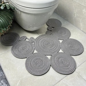 Puede incluir: Una alfombra de baño gris hecha de cuerda trenzada en forma circular. La alfombra está compuesta por varios círculos de diferentes tamaños.