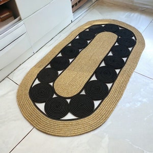 May include: An oval-shaped rug with a woven design. The rug features a tan border and a central section with black circular accents. The rug is placed on a white tile floor.
