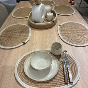 Op de afbeelding: Een set van vier ovale geweven placemats met een witte rand. De placemats zijn gemaakt van natuurlijke vezels en hebben een rustieke uitstraling. Een witte keramische kom en een witte keramische plaat liggen op een van de placemats. Een tafel van licht hout is op de achtergrond.