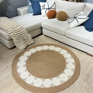 Puede incluir: Una alfombra redonda de fibra natural con un borde de cuerda blanca. La alfombra tiene un diseño tejido de círculos dentro de círculos. La alfombra está sobre un suelo de madera clara delante de un sofá seccional blanco.