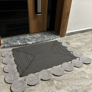 May include: A gray rectangular doormat with a decorative border of gray rope circles. The doormat is made of a woven material and has a textured surface.