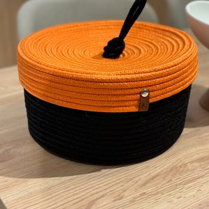 Puede incluir: Una cesta de almacenamiento redonda, naranja y negra con un asa de cuerda. La cesta est&aacute; hecha de cuerda trenzada y tiene una tapa.