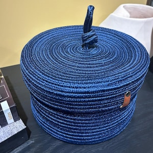 Puede incluir: Una cesta de almacenamiento azul tejida con tapa y asa de cuero. La cesta est&aacute; hecha de un material grueso parecido a una cuerda y tiene una forma circular.