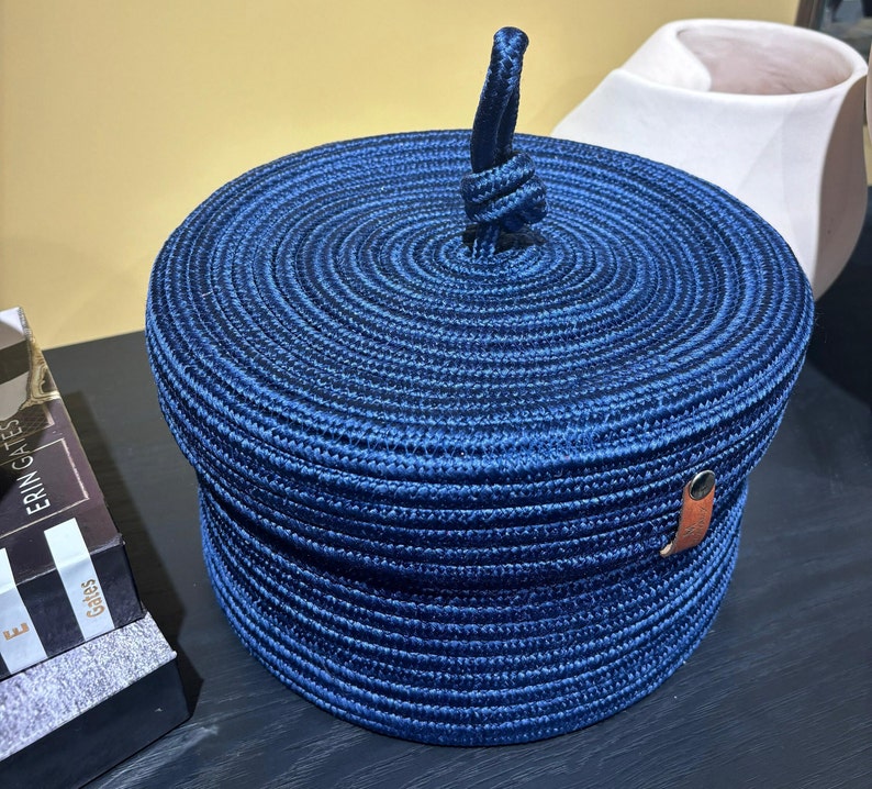 Puede incluir: Una cesta de almacenamiento azul tejida con tapa y asa de cuero. La cesta est&aacute; hecha de un material grueso parecido a una cuerda y tiene una forma circular.