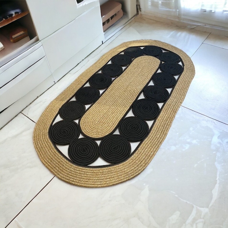 May include: An oval-shaped rug with a woven design. The rug features a tan border and a central section with black circular accents. The rug is placed on a white tile floor.