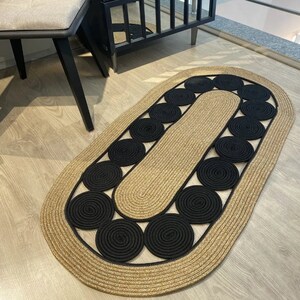 May include: Oval jute rug with a tan border and a central design of black circular accents. The rug's design features a repeating pattern of black circles, adding a modern touch to the floor covering. The rug is suitable for indoor use.
