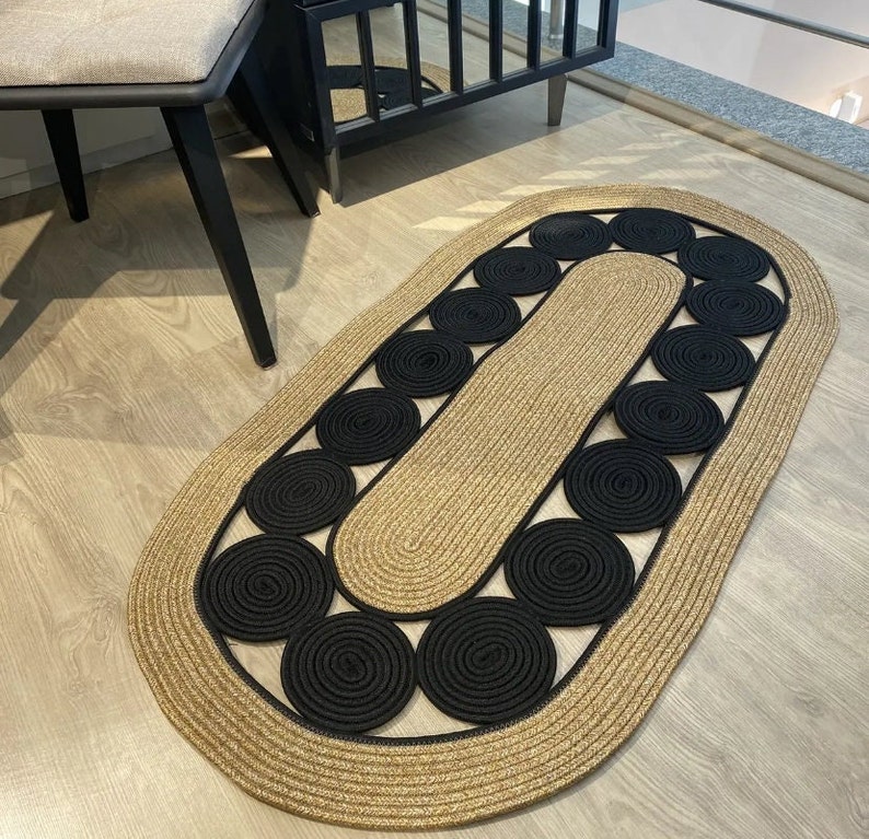 May include: Oval jute rug with a tan border and a central design of black circular accents. The rug's design features a repeating pattern of black circles, adding a modern touch to the floor covering. The rug is suitable for indoor use.