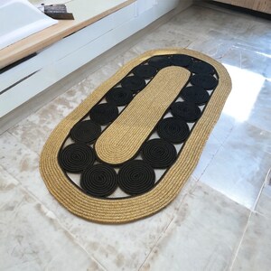 May include: Oval-shaped rug with a gold border and a central design of black circular patterns. The rug's interior features a gold-colored section, creating a contrasting aesthetic. The rug is made of woven material.