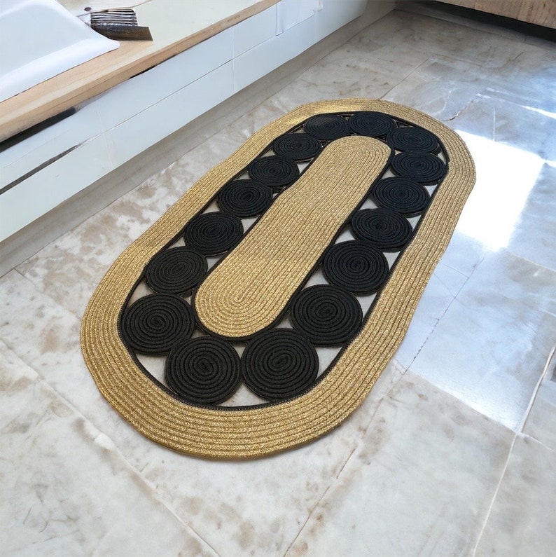 May include: Oval-shaped rug with a gold border and a central design of black circular patterns. The rug's interior features a gold-colored section, creating a contrasting aesthetic. The rug is made of woven material.