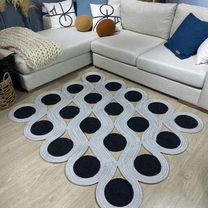 May include: A modern area rug with a unique design. The rug features a pattern of interconnected, wavy lines in a light gray color, with large, circular black accents. The rug is placed on a light-colored wooden floor, with a sectional sofa and decorative pillows in the background.