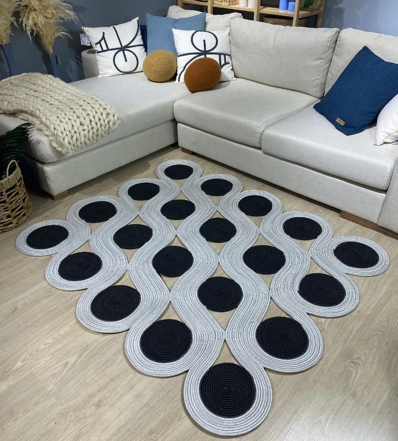 May include: A modern area rug with a unique design. The rug features a pattern of interconnected, wavy lines in a light gray color, with large, circular black accents. The rug is placed on a light-colored wooden floor, with a sectional sofa and decorative pillows in the background.