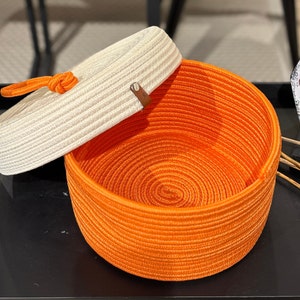 Puede incluir: Una cesta de almacenamiento tejida de color blanco y naranja con tapa. La tapa est&aacute; hecha de un material tejido y tiene una correa de cuero marr&oacute;n.