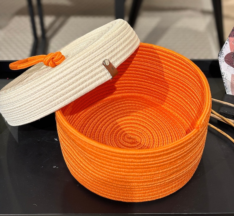 Puede incluir: Una cesta de almacenamiento tejida de color blanco y naranja con tapa. La tapa est&aacute; hecha de un material tejido y tiene una correa de cuero marr&oacute;n.