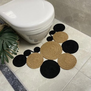 May include: A bathroom rug made of woven rope in black and gold circles. The rug is placed in front of a white toilet.