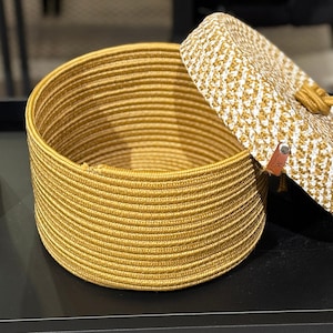 May include: A woven storage basket with a lid. The basket is made of natural fibers and has a light brown color. The lid is white with a brown trim.
