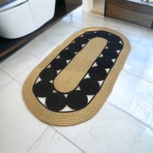 May include: Oval jute rug with a gold border and black circular accents. The rug has a central oval section and is surrounded by a border of tightly woven jute. The black accents are evenly spaced around the central section.