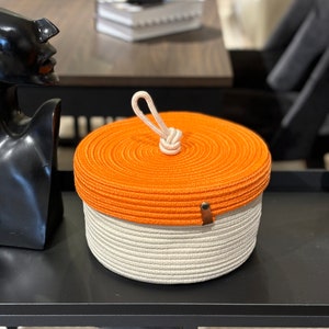 Puede incluir: Cesta de almacenamiento redonda tejida en naranja y blanco con asa de cuerda y tapa. La cesta es redonda y tiene una textura tejida.