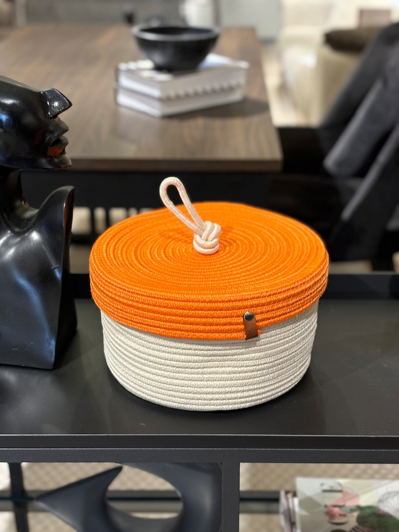 Puede incluir: Cesta de almacenamiento redonda tejida en naranja y blanco con asa de cuerda y tapa. La cesta es redonda y tiene una textura tejida.