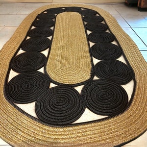 May include: An oval jute rug with a unique design. The rug features a central section and border in a natural tan color, with a series of black circular accents. The rug's design adds a touch of modern style to any room.