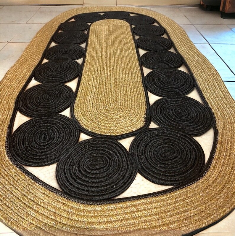 May include: An oval jute rug with a unique design. The rug features a central section and border in a natural tan color, with a series of black circular accents. The rug's design adds a touch of modern style to any room.