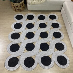 May include: A modern area rug with a repeating pattern of gray wavy lines and black circular accents. The rug is made of a woven material and is placed on a light-colored wooden floor. The design is geometric and contemporary.