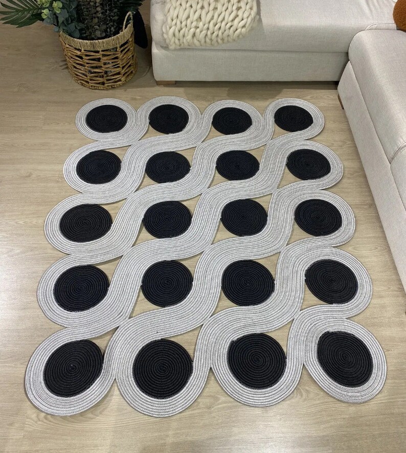 May include: A modern area rug with a repeating pattern of gray wavy lines and black circular accents. The rug is made of a woven material and is placed on a light-colored wooden floor. The design is geometric and contemporary.