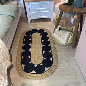 May include: Oval jute rug with a natural tan border and center, accented with a pattern of black circles. The rug is placed on a light wood floor, suggesting a home decor setting. The design is simple yet stylish, suitable for various interior styles.