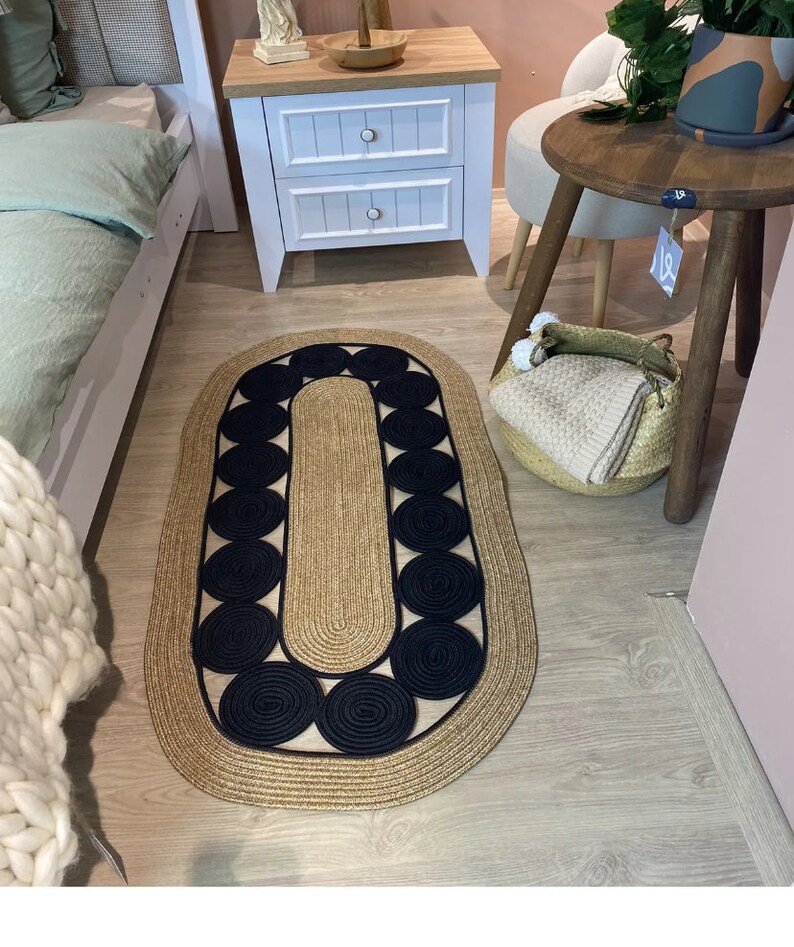 May include: Oval jute rug with a natural tan border and center, accented with a pattern of black circles. The rug is placed on a light wood floor, suggesting a home decor setting. The design is simple yet stylish, suitable for various interior styles.