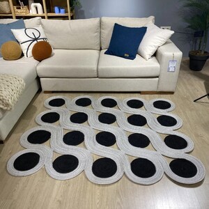 May include: A modern area rug with a unique design. The rug features a wavy pattern in light gray with large, circular black accents. The rug is placed on a light wood floor in front of a beige sofa with decorative pillows.