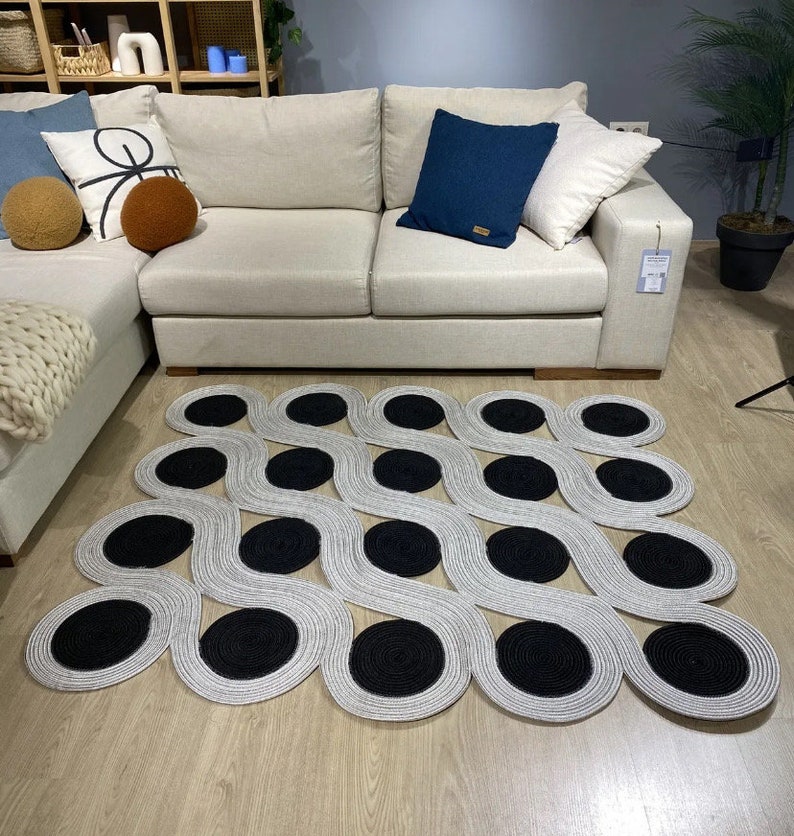May include: A modern area rug with a unique design. The rug features a wavy pattern in light gray with large, circular black accents. The rug is placed on a light wood floor in front of a beige sofa with decorative pillows.
