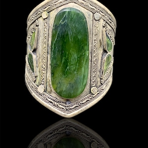 Può includere: Un bracciale a polsino argentato con una grande pietra verde ovale al centro. Il bracciale presenta dettagli intricati e pietre verdi più piccole sui lati. Il design è simmetrico e il bracciale è su sfondo nero.