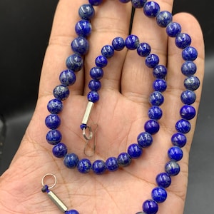 Puede incluir: Un collar con cuentas de lapislázuli azul oscuro. Las cuentas redondas tienen un acabado pulido y están ensartadas con un cierre plateado. El collar se muestra sobre un fondo neutro, resaltando el color vibrante de las piedras.