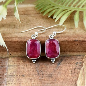 May include: A pair of drop earrings featuring rectangular, faceted deep red ruby gemstones set in polished silver frames. Each earring hangs from a simple silver hook wire, with a small decorative silver bead detail at the base of the stone setting.