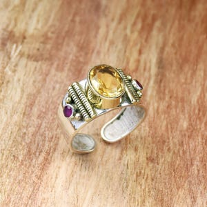 May include: Silver ring with a large, oval, yellow gemstone at the centre. The ring has gold-tone detailing and two small, round, red gemstones. The band is open at the base.