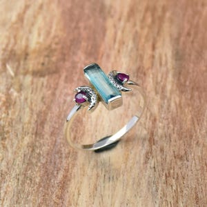 Puede incluir: Un anillo de plata con una piedra rectangular azul claro en el centro, flanqueada por dos pequeñas piedras rojas redondas. El anillo tiene un diseño único y moderno con elementos decorativos.
