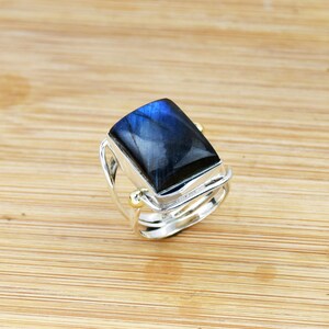 May include: A silver ring with a large, rectangular, dark blue gemstone. The ring has a double band design with small gold accents. The gemstone has a reflective, iridescent quality, with shades of blue and black.