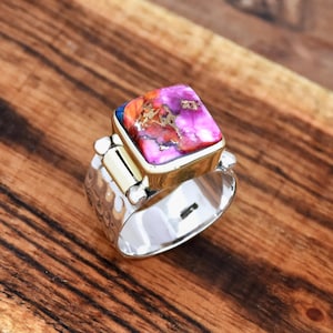 May include: A silver ring with a square-shaped pink, orange, and brown gemstone set in a gold setting. The ring has a textured band.
