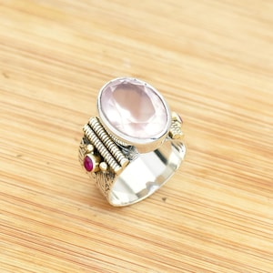 Pode incluir: Anel de prata com uma grande pedra oval rosa claro. O anel tem detalhes dourados e duas pequenas pedras vermelhas. Apresentado sobre uma superfície de madeira castanha clara.
